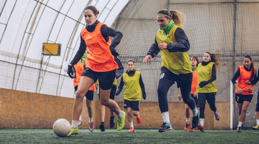 Female soccer players wearing thermal gear and bibs engage in an intense indoor practice session during winter.