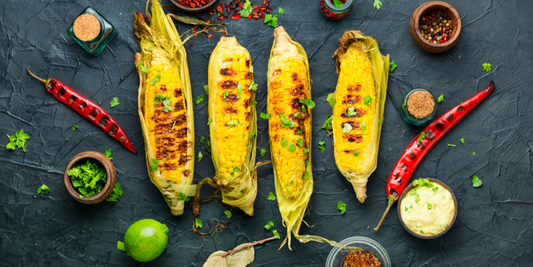 Cooked corn on the cob surrounded by lime wedges and garnishes on a dark wooden surface.