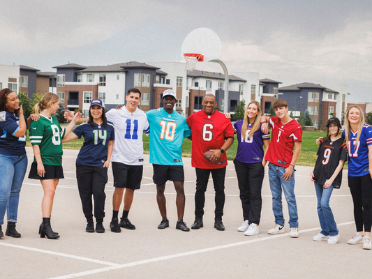 A group of athletes stands in a line on a basketball court, all wearing different sports jerseys. They smile, laugh, and lean on each other, capturing a moment of camaraderie. The backdrop includes a basketball hoop and residential buildings.