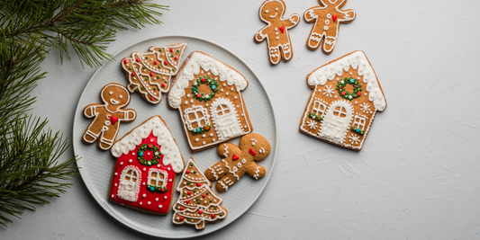 A plate of decorated gingerbread cookies featuring gingerbread men, trees, and houses with intricate icing designs, surrounded by greenery on a white background.