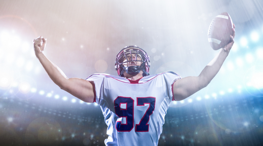 A football player in a white and red jersey celebrating under stadium lights, holding a football with arms raised triumphantly.