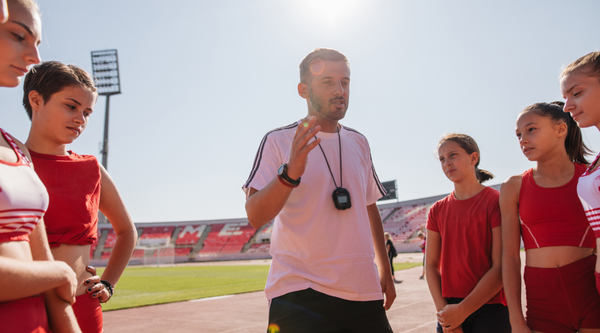 A coach stands in front of a group of young female athletes at a track, giving instructions. The athletes listen attentively, dressed in red training uniforms, with a bright sunlit stadium in the background.