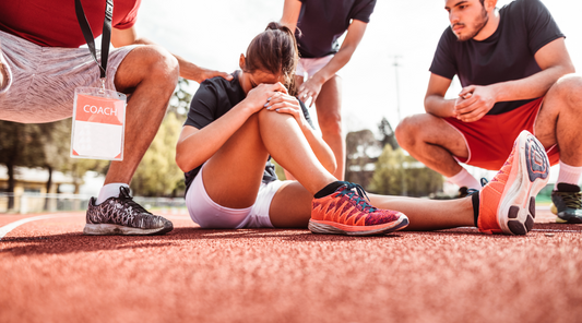 A female athlete sits on the track, head down in her hands, as teammates and a coach offer support after an injury.