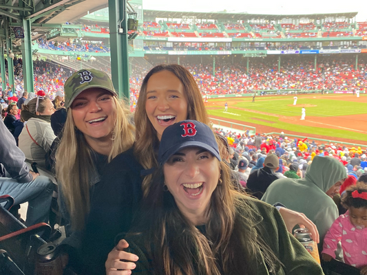 Three women, wearing Boston Red Sox hats, smile and laugh while attending a game at Fenway Park. The crowd around them is filled with excited fans as they enjoy the baseball game on a cool, overcast day.