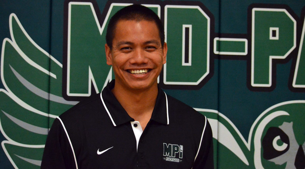 Coach Gabriel Maunupau, wearing a black athletic polo with "MP Athletics" embroidered on it, smiles in front of a green backdrop featuring a sports logo.