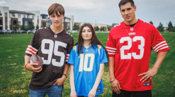 A group of athletes stands in a line on a basketball court, all wearing different sports jerseys. They smile, laugh, and lean on each other, capturing a moment of camaraderie. The backdrop includes a basketball hoop and residential buildings.