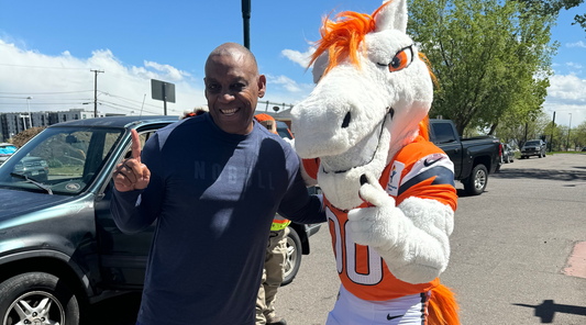 Mike posing outside in a Denver Broncos-themed setting, standing next to the Broncos mascot, smiling while making a number-one gesture.