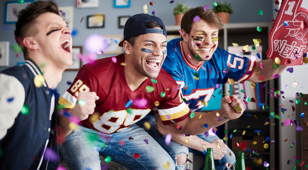 Three excited football fans, wearing jerseys and face paint, celebrate in a living room while confetti flies around them. They are shouting and cheering, clearly invested in a game they are watching on TV, with expressions of pure joy and excitement.