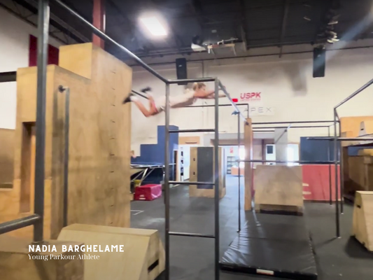 Nadia Barghelame, a young parkour athlete, is captured mid-air as she performs a dynamic parkour jump between structures in an indoor training gym.