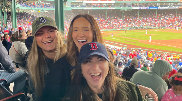 Three women, wearing Boston Red Sox hats, smile and laugh while attending a game at Fenway Park. The crowd around them is filled with excited fans as they enjoy the baseball game on a cool, overcast day.