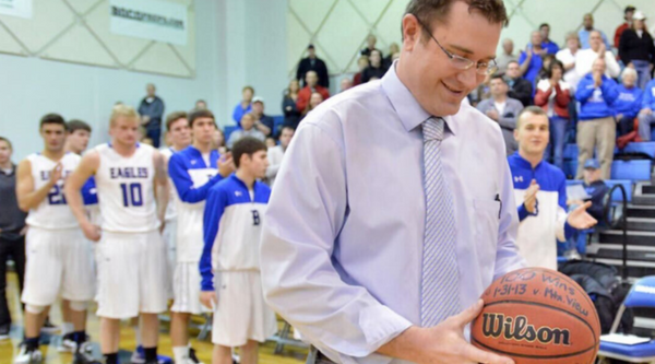 Coach Terrence Dunn looks down at a commemorative basketball in his hands, smiling. He is surrounded by his Eagles team members, who stand in the background, watching and applauding the achievement.