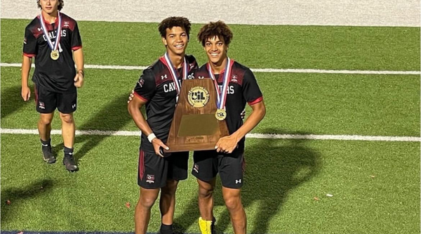 Tre and Skyler Wright smiling and holding a large trophy, both wearing black and red soccer uniforms and medals, standing on a green field after winning the state championship.