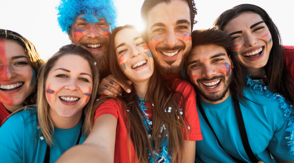 A group of smiling fans, all wearing face paint and brightly colored team shirts, take a joyful selfie together. The sun shines behind them, adding a warm glow to the group’s cheerful faces, full of team spirit and camaraderie.