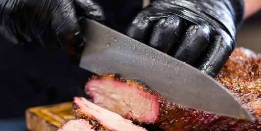 Close-up of a gloved hand using a knife to carve a smoked brisket on a wooden cutting board.