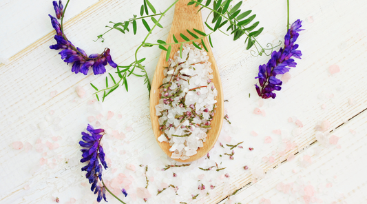 A wooden spoon filled with Epsom salts, surrounded by purple flowers on a white background.